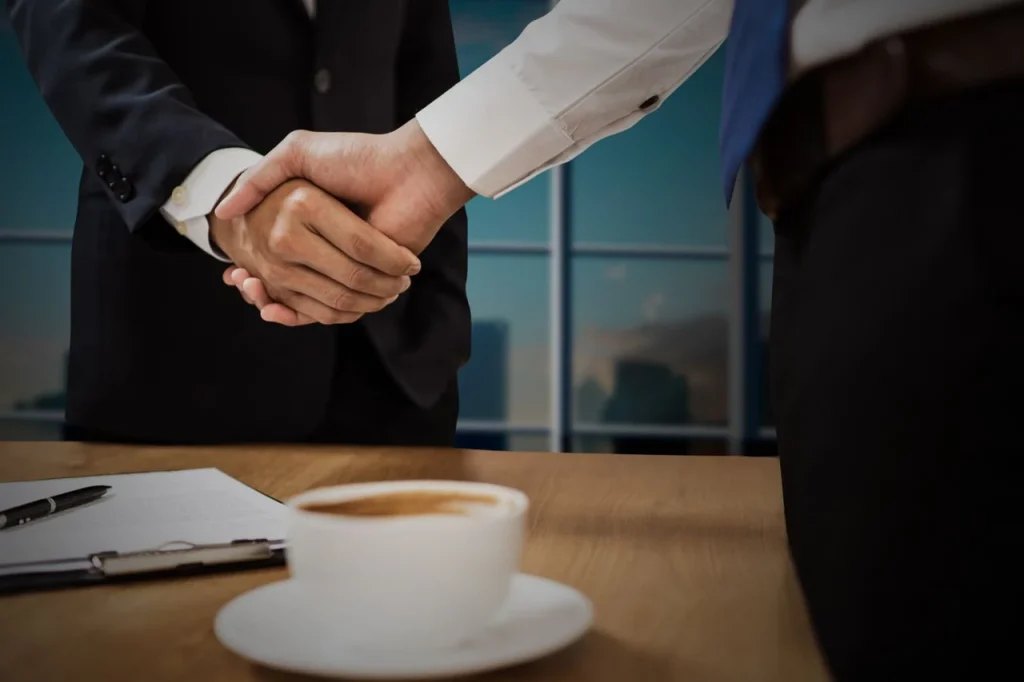 Negociação entre empresas no e-commerce B2B: dois profissionais de terno fazem aperto de mãos em reunião, com mesa e xícaras ao fundo, simbolizando acordos comerciais.