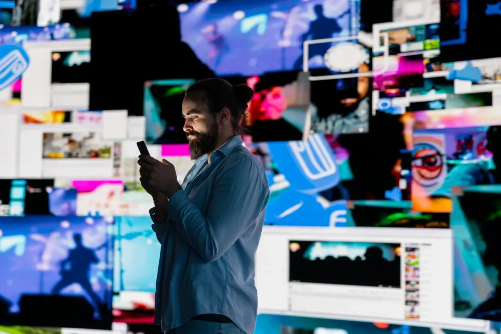 O Renascimento do colecionável físico na era do Digital-First: O Que Isso Diz Sobre a Saturação de Telas: Homem em frente a monitores com gráficos e imagens coloridas, usando o celular em um ambiente de tecnologia e mídia digital, representando a ideia de Digital-First e saturação de telas