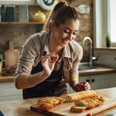 Young happy woman making bruschetta while preparing food in the kitchen. Poucos alimentos carregam tanta fama negativa quanto o pãozinho de todo dia. Foto: freepik
