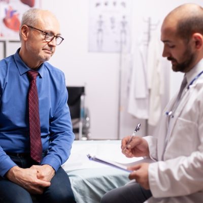 Surgeon wearing stethoscope discussing treatment in examination room with senior man. Elderly patient having a converstation with medical stuff in private clinic during consultation.