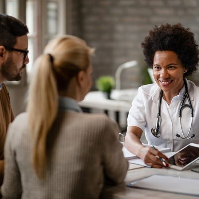 Happy black female doctor talking to a couple and showing them medical test results on a touchpad during consultations at clinic.
