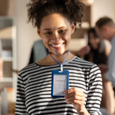 Jovem sorrindo em evento corporativo, segurando uma Credencial com cordão: Vantagens e aplicações para eventos, empresas e instituições, com crachá azul preso ao pescoço.