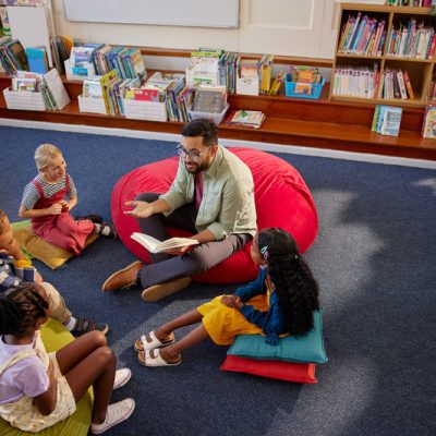 High angle view of enthusiastic male teacher reading a book to primary students while sitting in school library with copy space. Curious elementary schoolchildren sitting on big cushion in library while listening to story from librarian. Top view of middle eastern man at primary school reading fairy tale during recess.