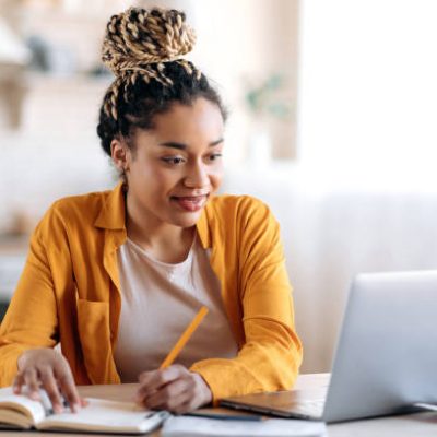 Focused cute stylish african american female student with afro dreadlocks, studying remotely from home, using a laptop, taking notes on notepad during online lesson, e-learning concept, smiling