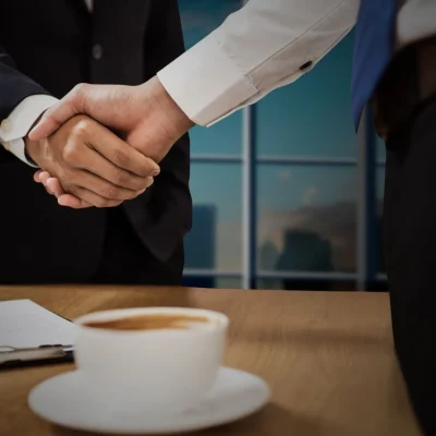 Negociação entre empresas no e-commerce B2B: dois profissionais de terno fazem aperto de mãos em reunião, com mesa e xícaras ao fundo, simbolizando acordos comerciais.