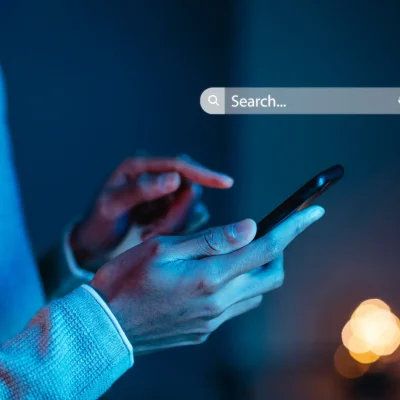 Pessoa usando um smartphone em ambiente com iluminação baixa, com foco na tela do dispositivo e uma barra de pesquisa do Google na parte superior da imagem, simbolizando estratégias de SEO na prática para aparecer no Google.