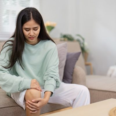A young woman sits on a couch, holding her knee in discomfort, highlighting her focus on health awareness and the importance of self-care. Dores nas articulações podem ir além do desgaste do dia a dia e indicar doenças autoimunes, como lúpus e artrite reumatoide
Divulgação / Envato